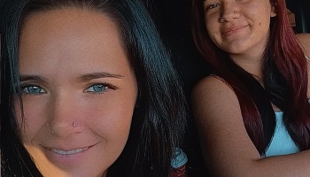 Close-up selfie of two smiling young women with nose piercings inside a vehicle.