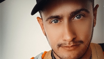 Close-up portrait of a young man with blue eyes, light facial hair, wearing a black cap and an orange safety vest.