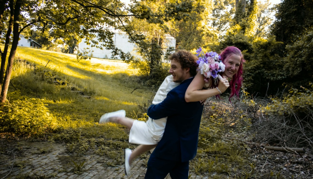 A smiling groom in a suit carries his bride with pink hair and a bouquet on a sunny, grassy field.