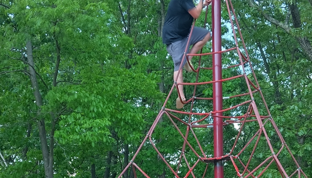 A man climbing a tall red rope pyramid playground structure surrounded by green trees.