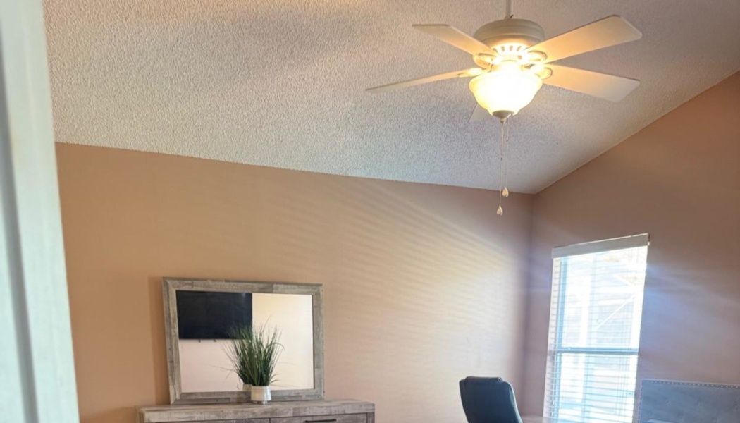 Bright bedroom with peach walls, a lighted ceiling fan, a wooden dresser with a mirror, and a sunlit window.