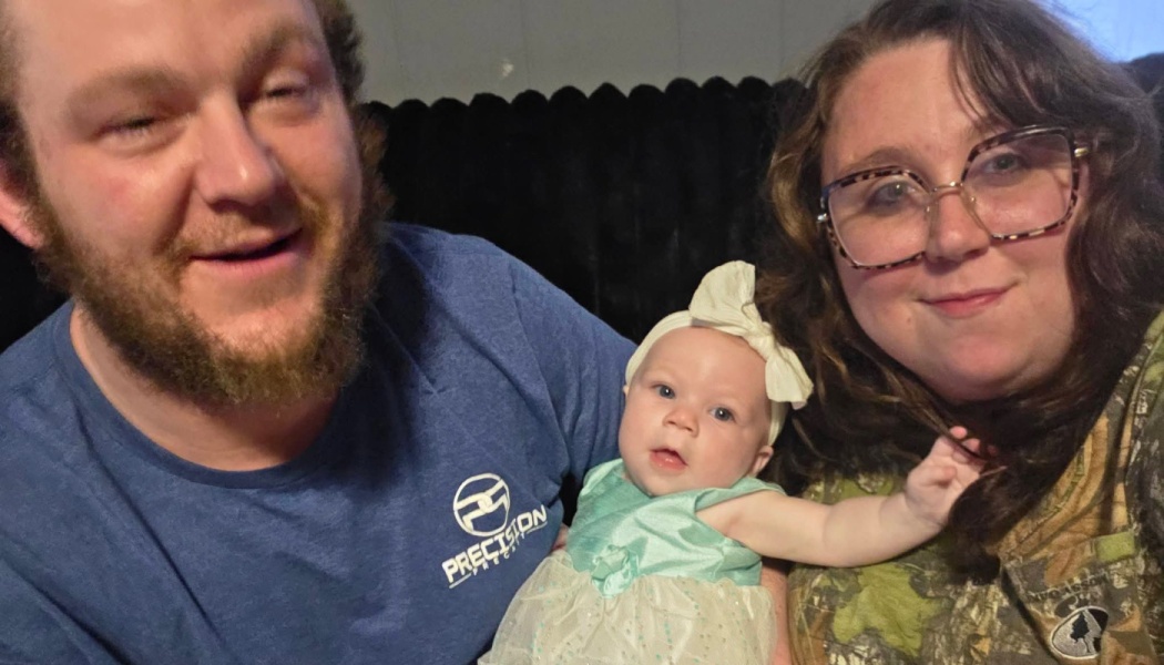 A smiling mother and father pose closely with their baby daughter wearing a mint green dress and white bow.