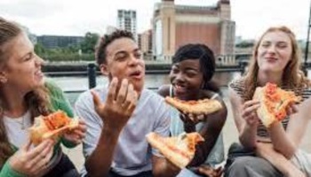 Four young friends smiling and eating pizza together outdoors near a city waterfront.