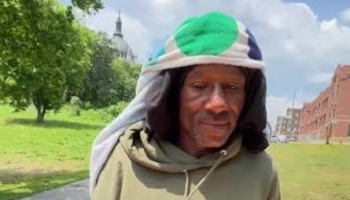 A man in an olive green hoodie with a colorful patterned head covering stands outdoors in a park.
