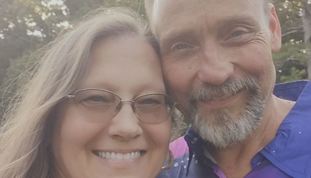 A smiling woman with glasses and a man with a grey goatee posing closely together outdoors.