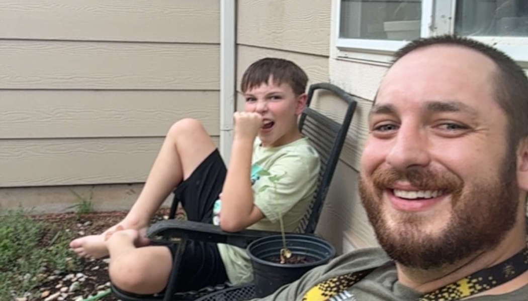 A smiling bearded man takes a selfie with a young boy making a playful face in a patio chair behind him.