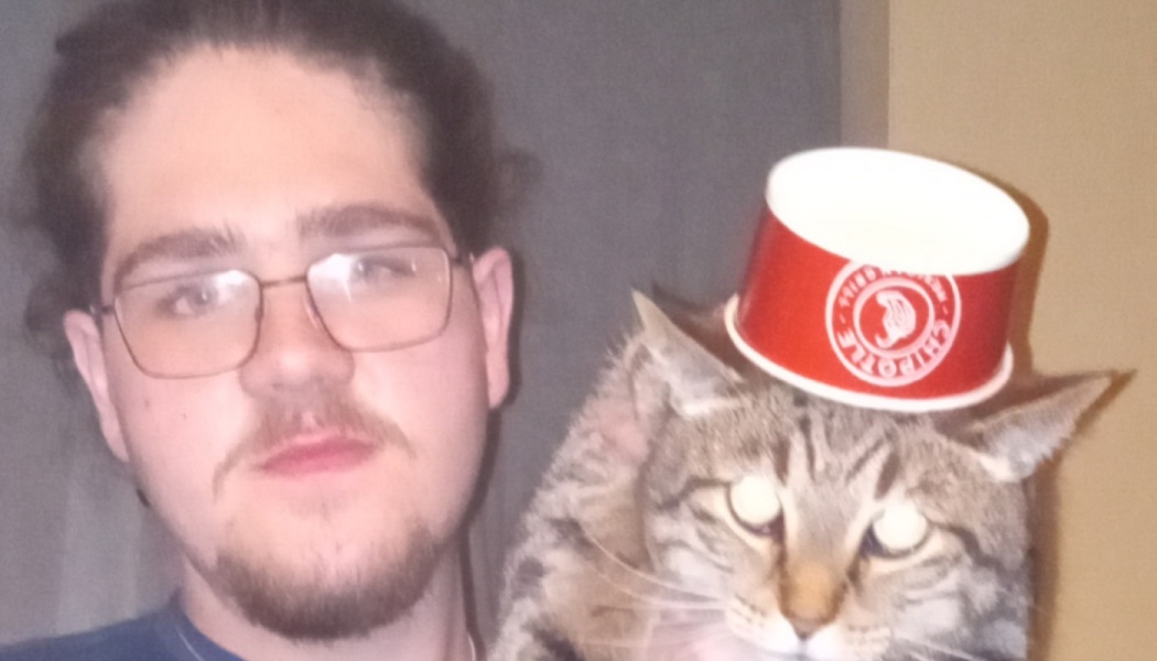 Man with glasses next to a tabby cat wearing a red Chipotle cup on its head like a hat.