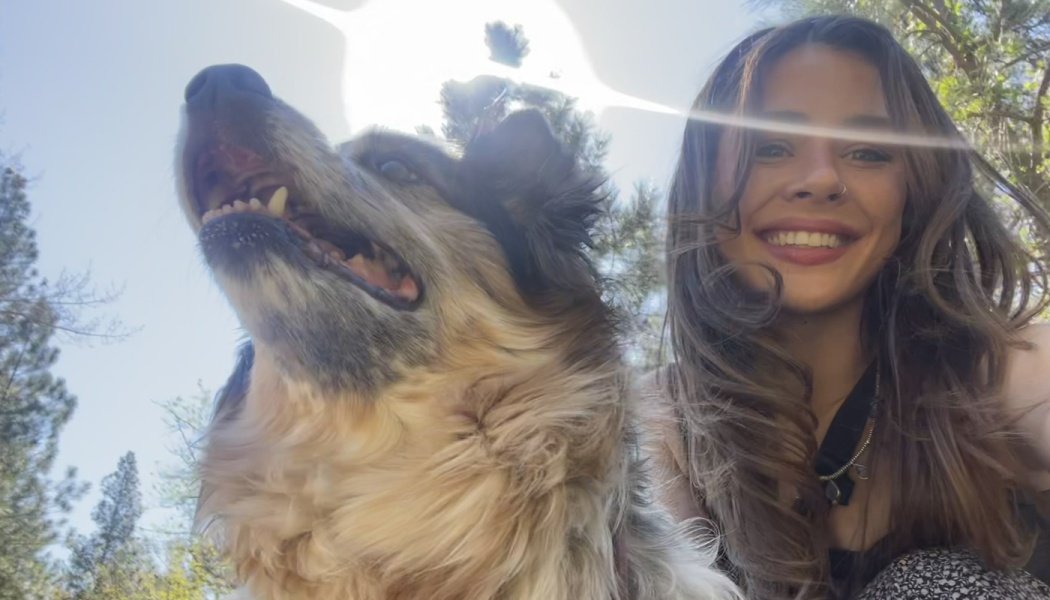 A smiling woman taking a sunny outdoor selfie with her happy, fluffy dog looking up.