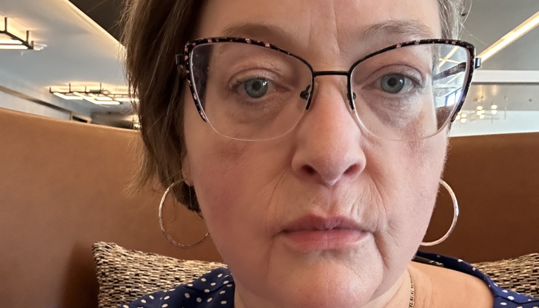 Close-up portrait of a woman with short hair, cat-eye glasses, hoop earrings, and a blue polka dot shirt.