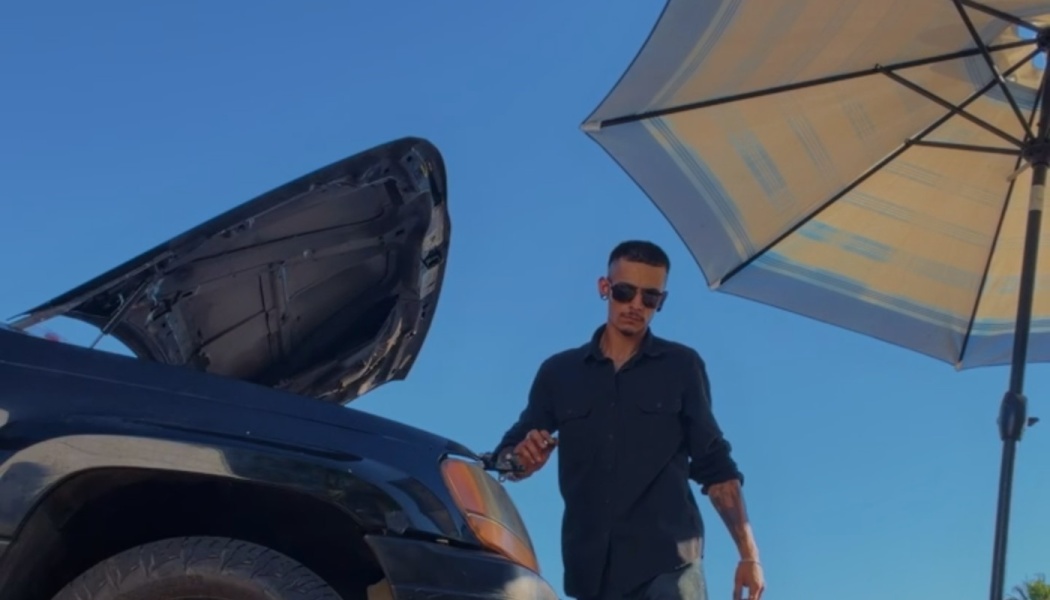 A man wearing sunglasses stands beside a black car with its hood open under a large sun umbrella.