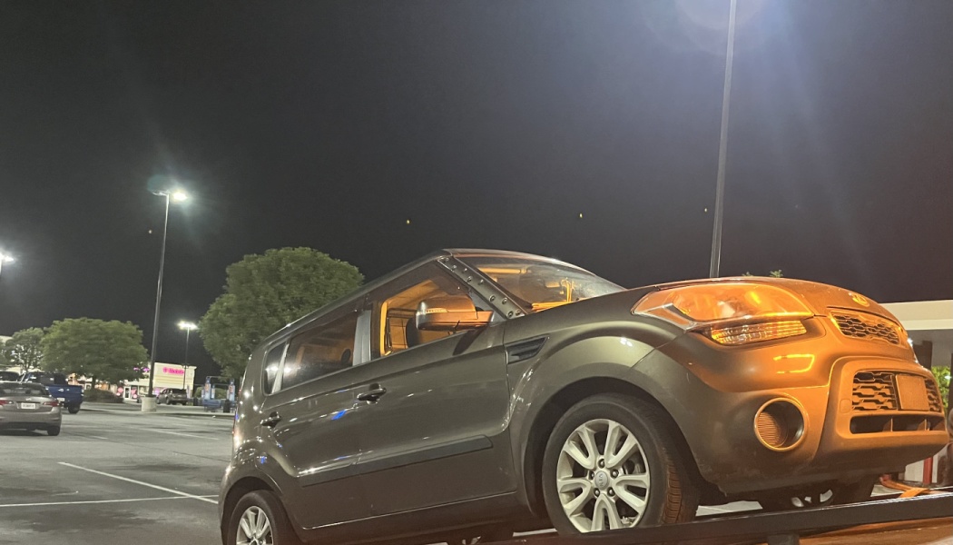 A grey Kia Soul sitting on the flatbed of a tow truck in a parking lot at night.