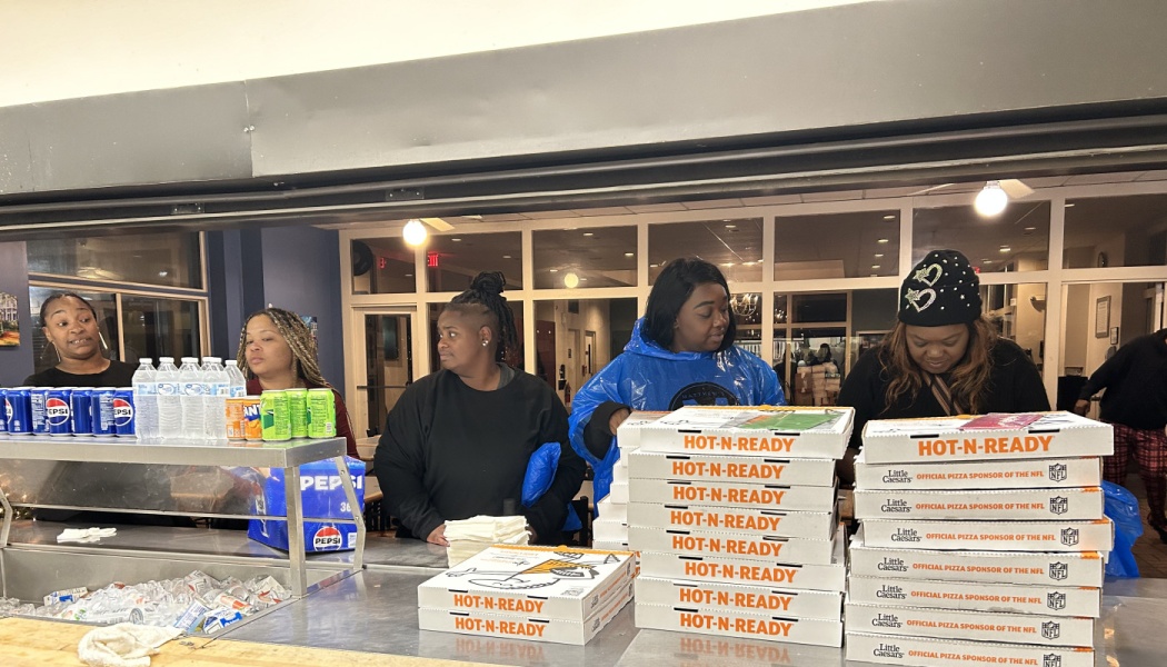 Volunteers stand behind a concession counter stacked with Little Caesars pizza boxes and drinks.