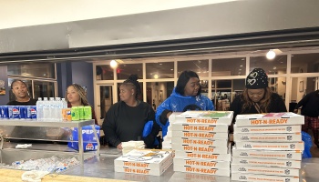 Volunteers stand behind a concession counter stacked with Little Caesars pizza boxes and drinks.