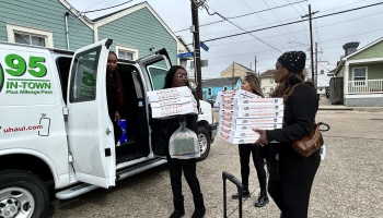 Women carry large stacks of pizza boxes from a white van on a residential street.