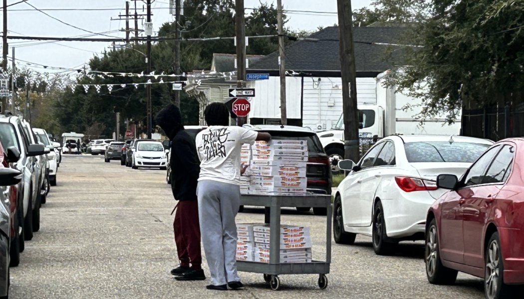Two people stand on a street by a utility cart stacked high with dozens of pizza boxes.
