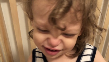 A toddler with curly hair scrunches their face and cries, wearing a blue and white striped shirt.
