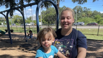 A smiling blonde woman holding a young toddler in a blue shirt at an outdoor playground near a swing set.