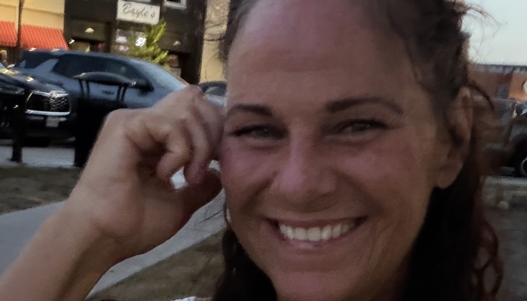 Close-up of a smiling woman with dark curly hair resting her hand near her head outdoors.