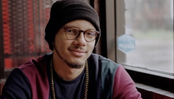 A smiling young man wearing round glasses, a black beanie, a gold chain, and a maroon sweatshirt near a window.