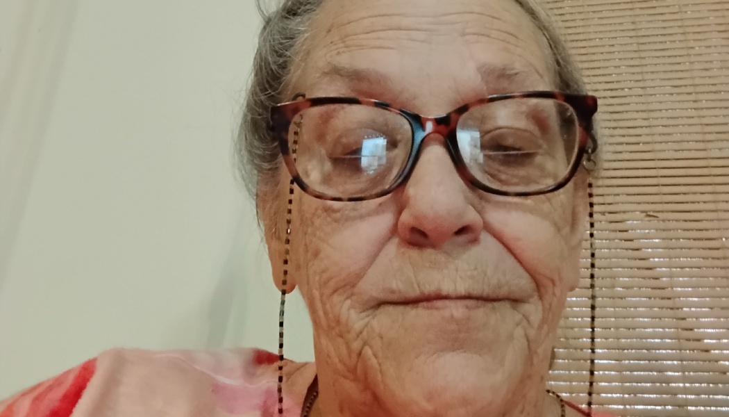 Close-up portrait of an elderly woman with grey hair, wearing tortoiseshell glasses attached to a beaded strap.