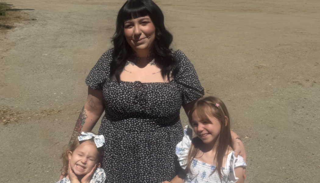 A mother in a dark floral dress smiling outdoors with her two young daughters.