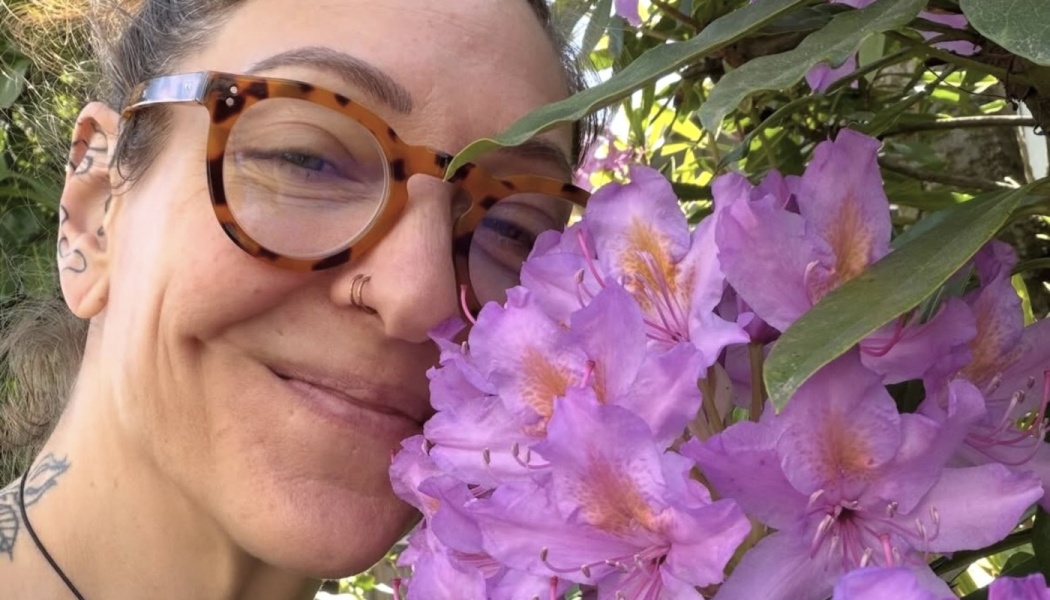 A smiling woman wearing tortoiseshell glasses poses close to a cluster of vibrant purple rhododendron flowers.
