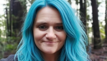 Close-up portrait of a woman with bright blue hair smiling outdoors in a forest.