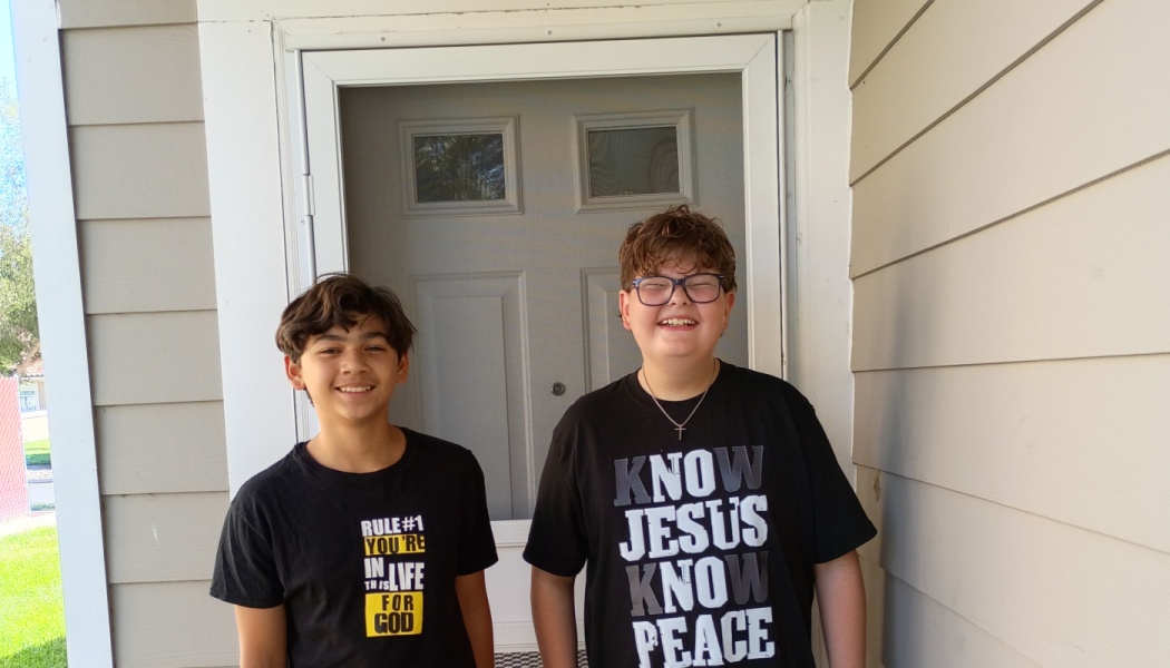 Two smiling young boys wearing black graphic t-shirts stand together on a front porch.