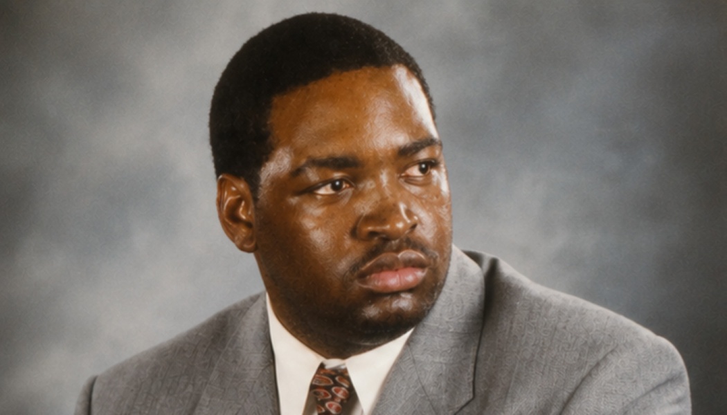 Headshot of a Black man in a grey suit and patterned tie looking to the side with a serious expression.