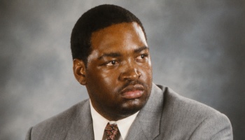 Headshot of a Black man in a grey suit and patterned tie looking to the side with a serious expression.