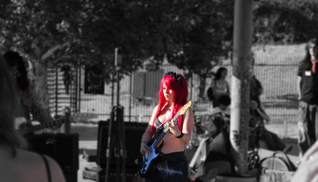 A musician with bright red hair plays electric guitar outdoors in a selective color photograph.