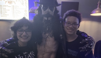 Two smiling teens pose with a performer in gothic face paint and a top hat at a music venue.