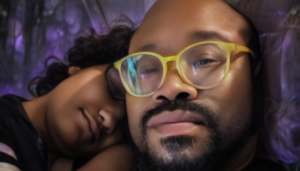 A man with yellow glasses and a beard lies down as a child rests their head on his shoulder.