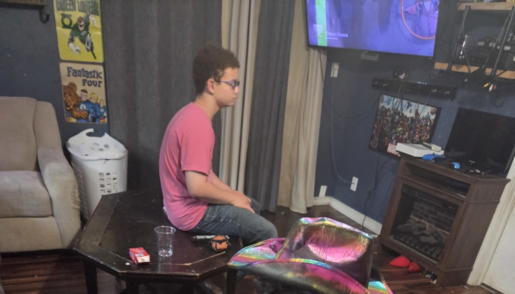 A young man in a pink shirt sits on a coffee table facing a television in a living room.