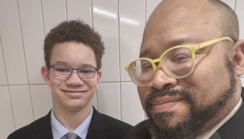 A smiling man with yellow glasses and a boy in a suit posing for a close-up selfie.
