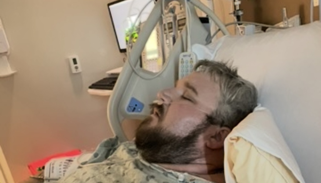 A bearded man with a nasal cannula rests with his eyes closed in a hospital bed.