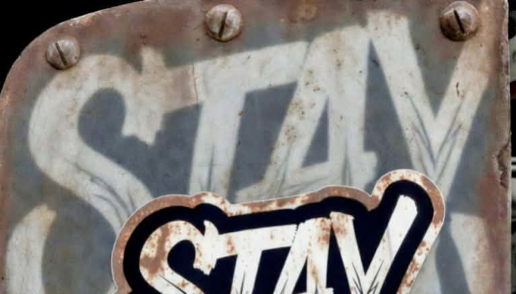 Stylized graffiti text spelling "STAY" on a rusty, weathered metal plate fastened with screws.
