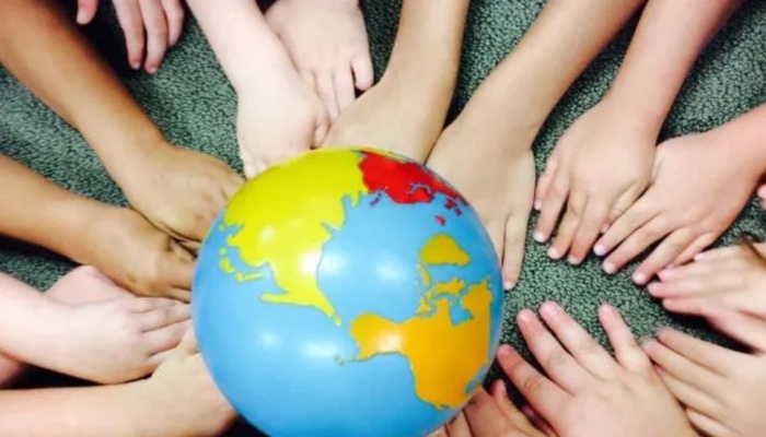 Children's hands of diverse skin tones placed in a circle around a colorful toy globe on a green carpet.