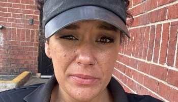 Close-up of a woman in a black visor and nose ring looking somberly at the camera against a red brick wall.