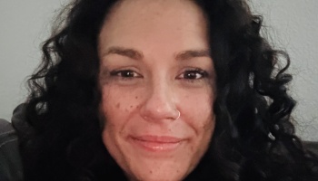 Close-up portrait of a smiling woman with dark curly hair and a nose ring.
