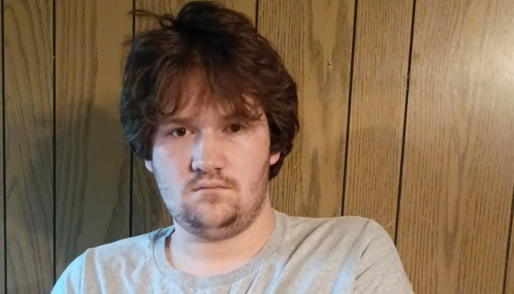 Young man with messy brown hair and scruff looking at the camera in front of wood wall paneling.
