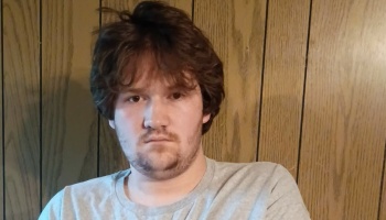 Young man with messy brown hair and scruff looking at the camera in front of wood wall paneling.