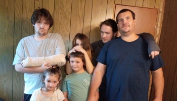 A family poses together indoors in front of a wood-paneled wall, including a man with a cast on his arm.