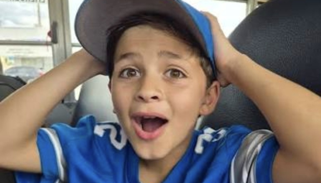 Young boy wearing a blue jersey and cap holds his head in excitement with a surprised, open-mouthed expression.