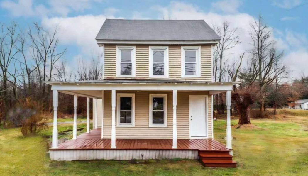 A two-story beige house with a white wraparound porch and reddish-brown wooden deck in a grassy yard.