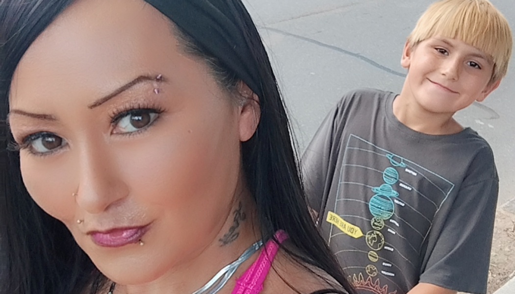 A woman with dark hair and facial piercings close-up, with a smiling blonde boy standing behind her.
