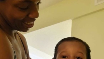 A smiling woman looks down lovingly at a young child looking towards the camera.