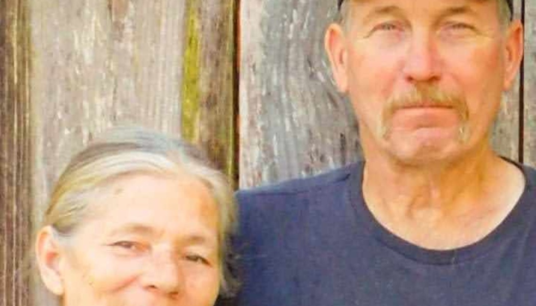 An older man with a mustache and a smiling gray-haired woman standing together in front of a rustic wood wall.