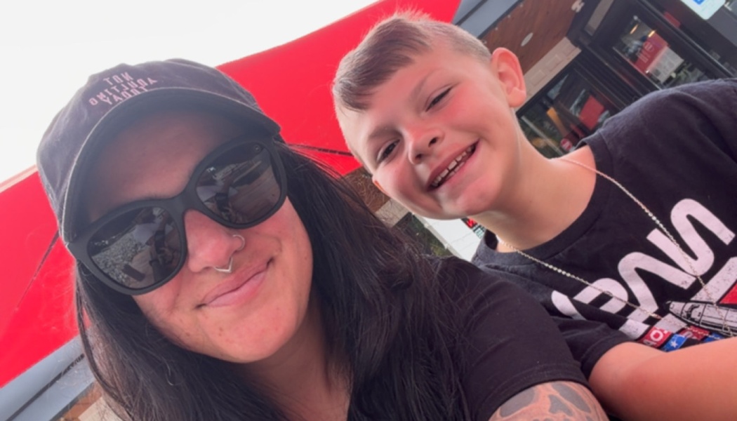 A smiling woman wearing dark sunglasses and a hat poses for a close selfie with a smiling young boy outdoors.