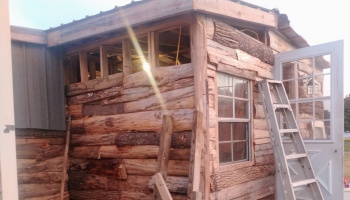 Rustic wooden cabin under construction with raw log siding, exposed framing, and a ladder leaning by the door.